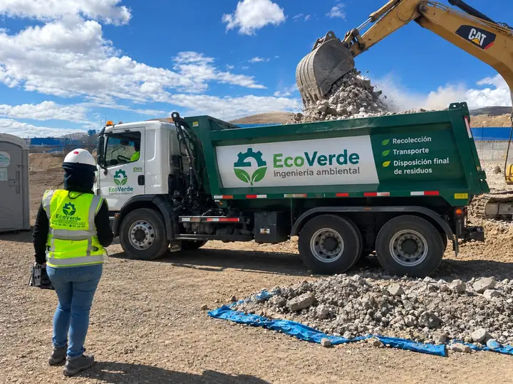 Camión de EcoVerde recibiendo escombros de una excavadora en un frente de trabajo, con una supervisora de la empresa observando la maniobra.