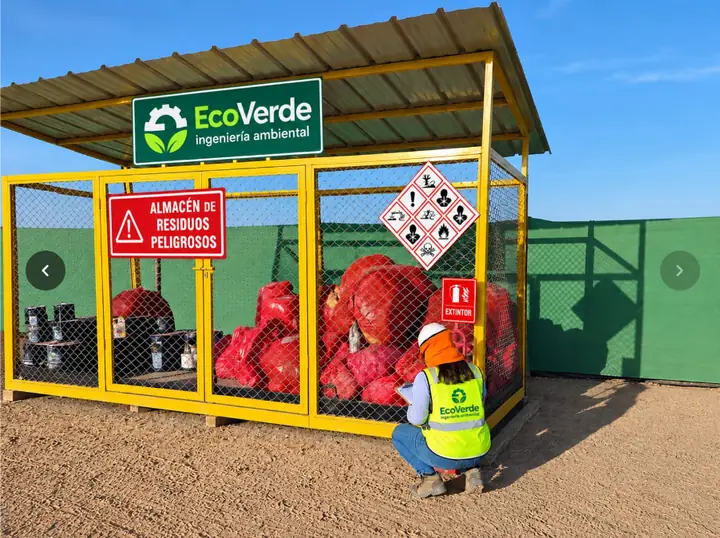 Almacén de residuos peligrosos de EcoVerde, con techo, cerco perimétrico, señalización de rombos MATPEL y una supervisora de la empresa registrando el inventario.